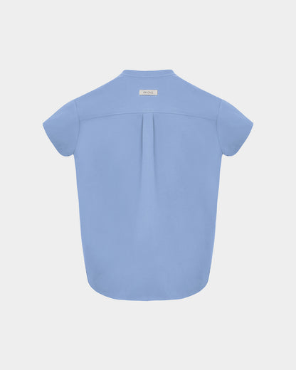 A light blue short-sleeve scrub shirt shown from the back, featuring a subtle center back pleat and a small rectangular brand label at the neckline, set against a solid light gray background.
