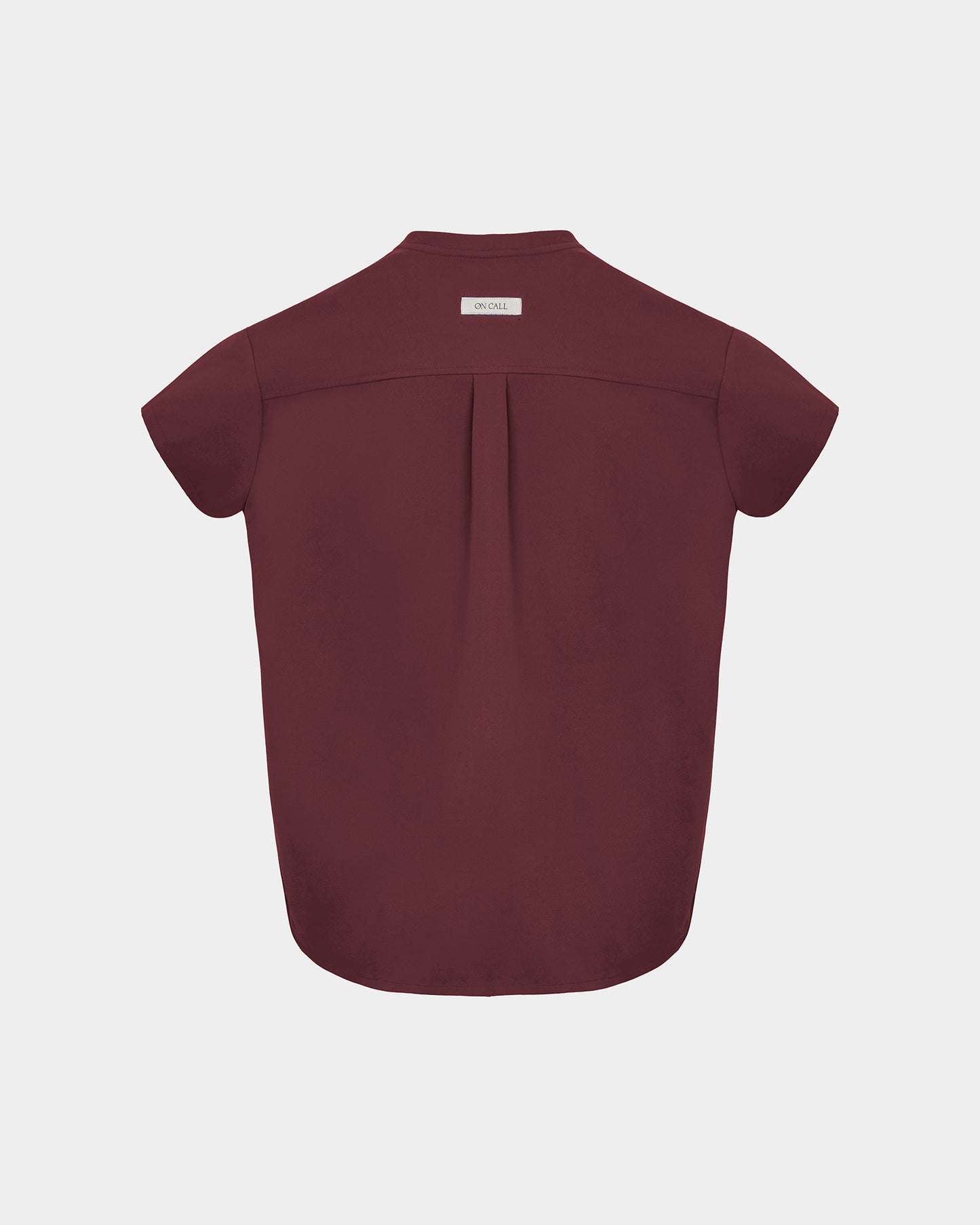 A short-sleeve scrub shirt shown from the back, featuring a subtle center back pleat and a small rectangular brand label at the collar, displayed on a light gray background.