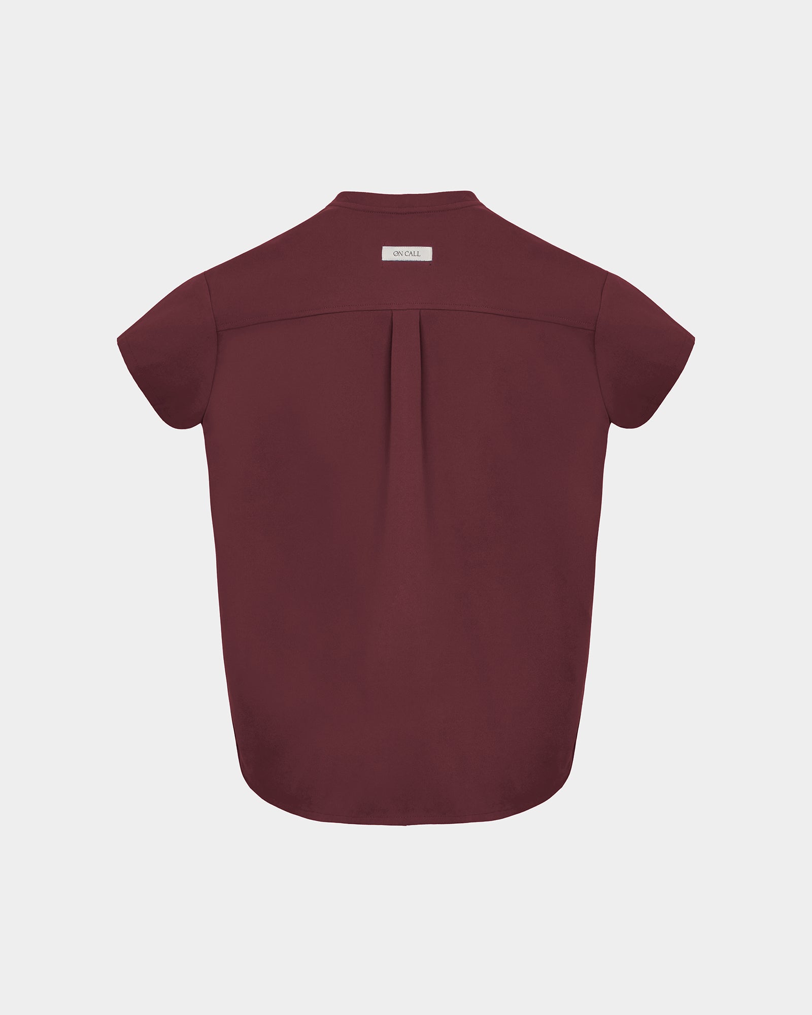 A short-sleeve scrub shirt shown from the back, featuring a subtle center back pleat and a small rectangular brand label at the collar, displayed on a light gray background.