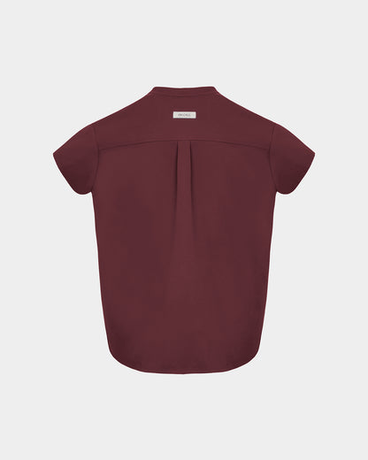 A short-sleeve scrub shirt shown from the back, featuring a subtle center back pleat and a small rectangular brand label at the collar, displayed on a light gray background.