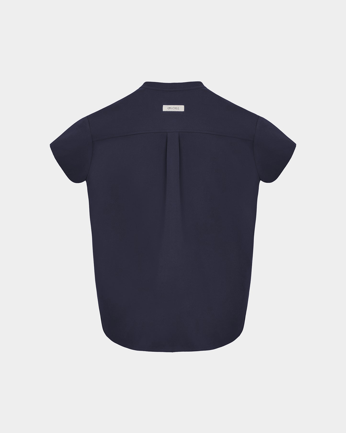 A navy short-sleeve scrub shirt shown from the back, featuring a subtle center back pleat and a small rectangular brand label at the collar, set against a light gray background.