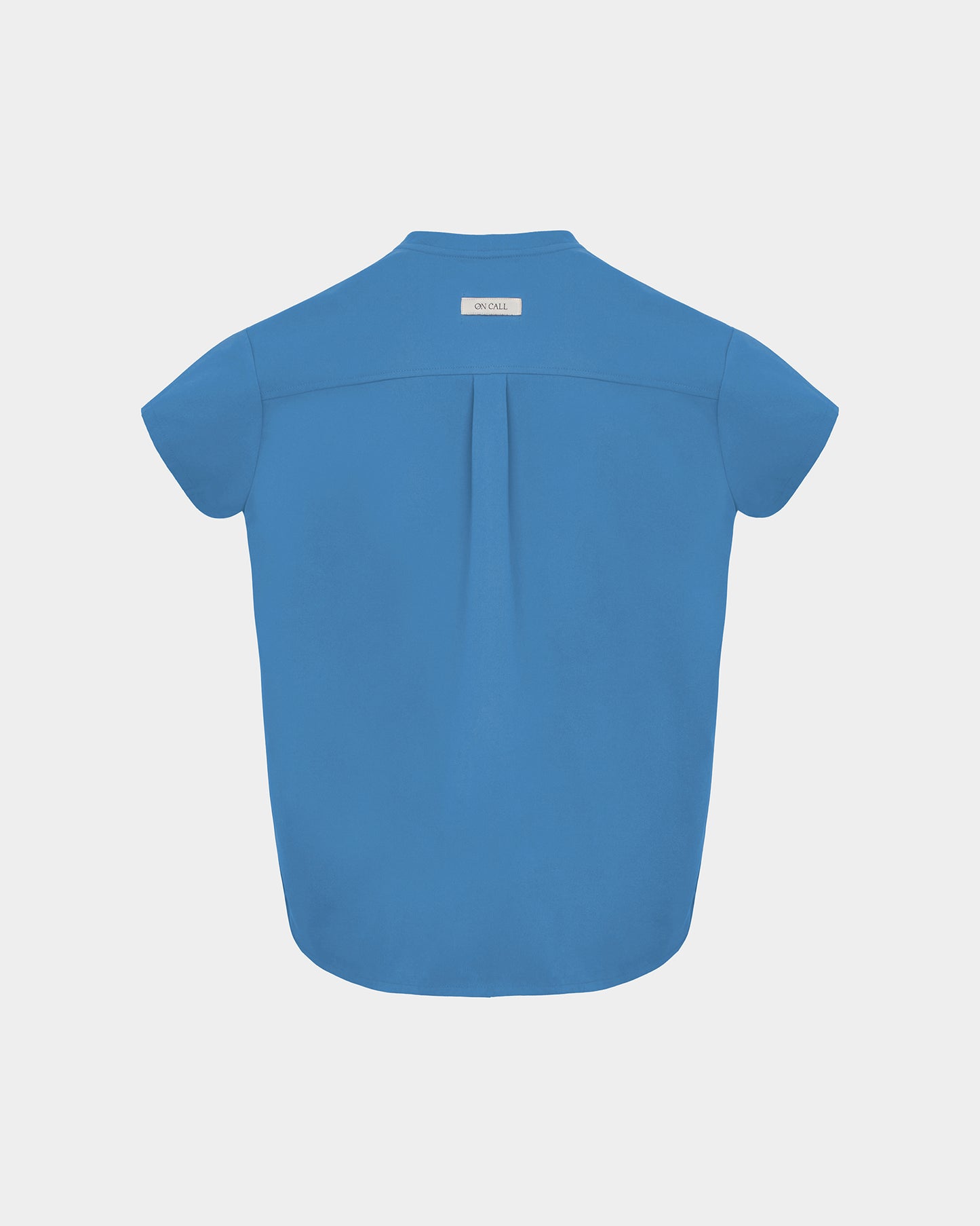 A short-sleeve bright blue scrub shirt shown from the back, featuring a subtle center back pleat and a small white brand label at the collar, set against a solid light gray background.