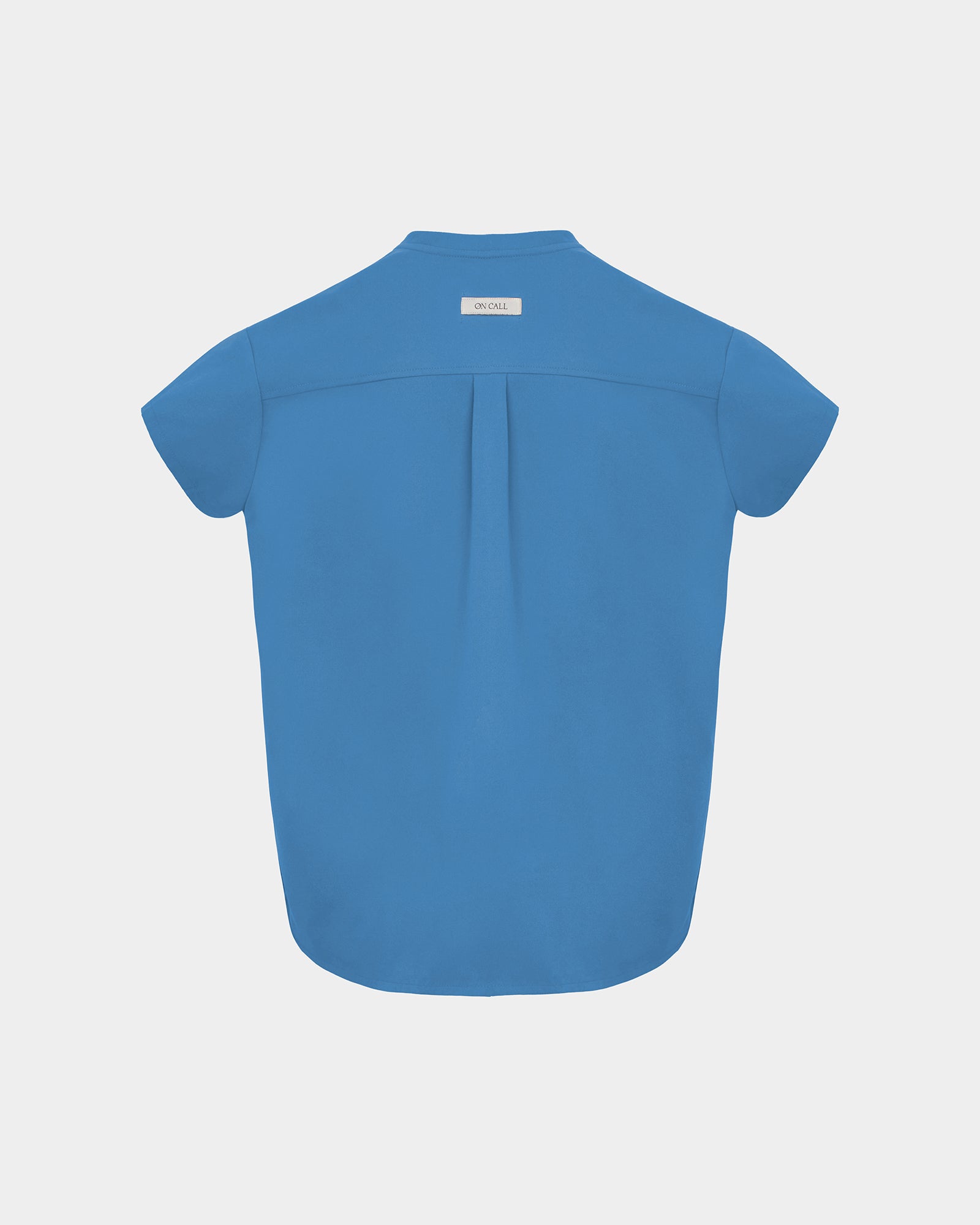 A short-sleeve bright blue scrub shirt shown from the back, featuring a subtle center back pleat and a small white brand label at the collar, set against a solid light gray background.