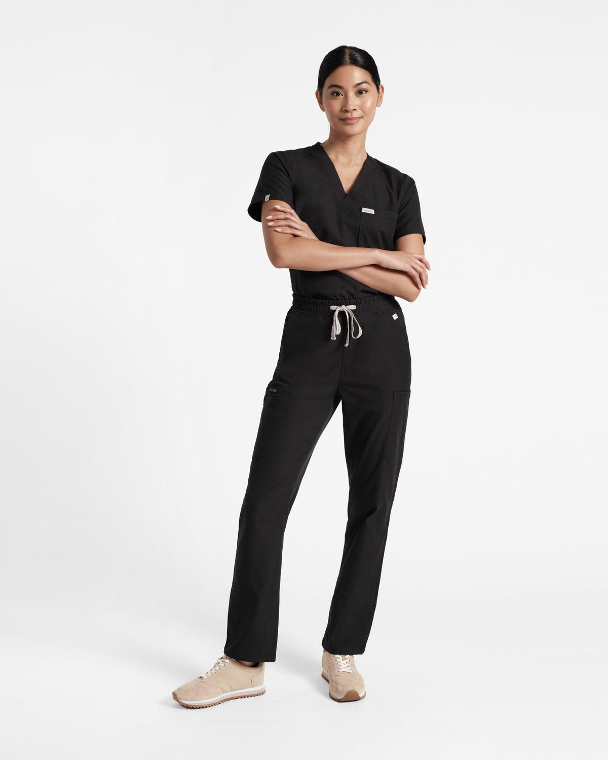 Woman wearing black v-neck scrub top with chest pocket tucked into black straight leg cargo scrub pants with arms crossed.
