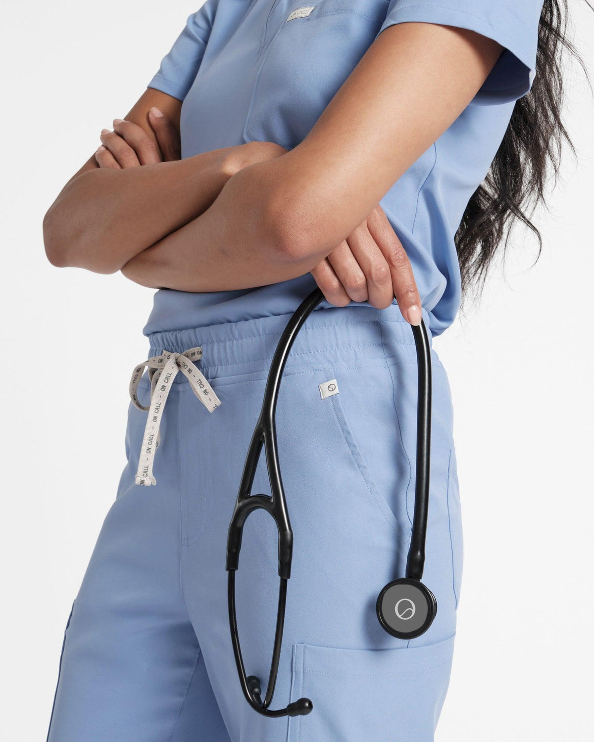 Woman in light blue cargo scrub pants and shirt with her arms crossed and holding a stethoscope. 