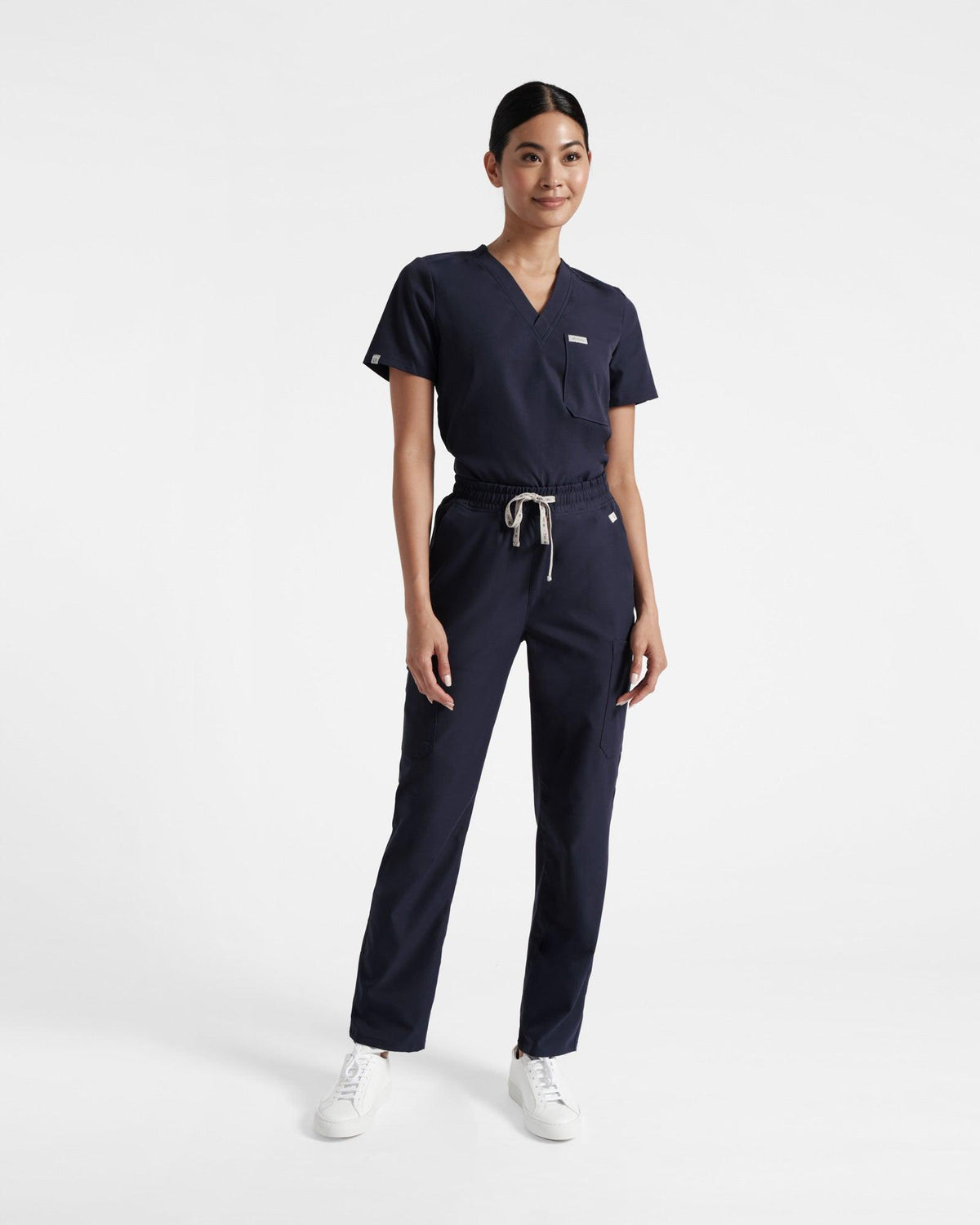 Woman in a navy v-neck scrub shirt tucked into matching straight leg cargo scrub pants. 