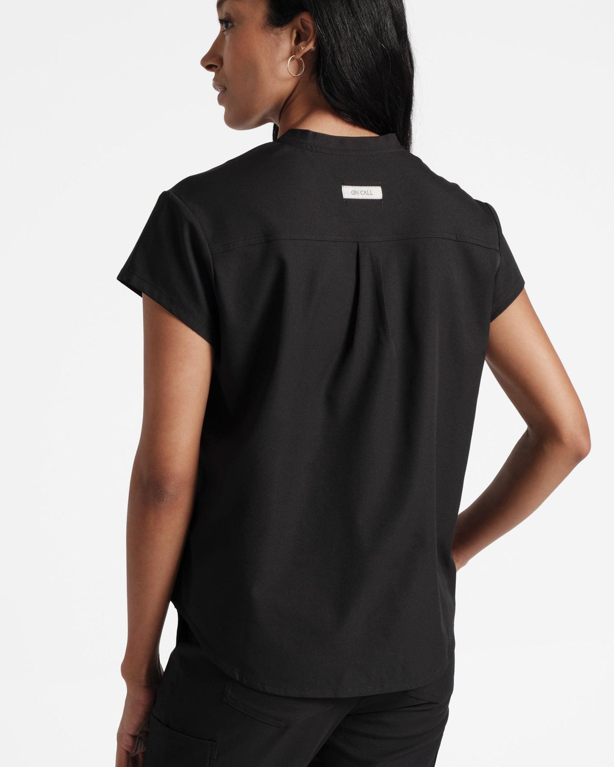 Back view of woman wearing black scrub top with a back pleat and branded On Call fabric label under neck seam.