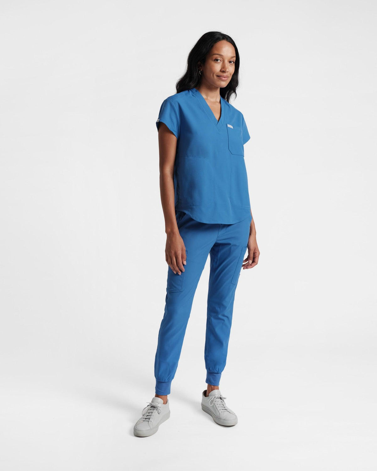 A woman wearing a v-neck, bright blue scrub top with a spacious chest pocket and a side pockets and matching cargo jogger scrub pants.