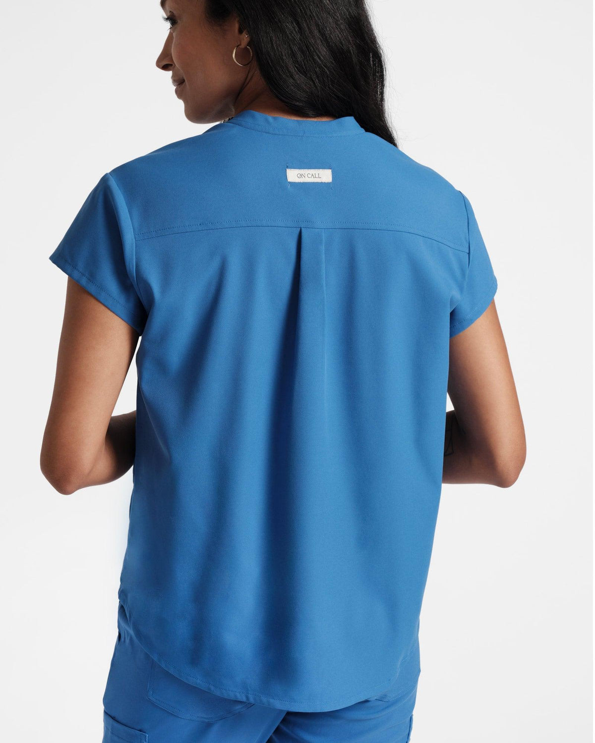 Back view of woman wearing bright blue scrub shirt with a back pleat and On Call fabric label on the yoke.