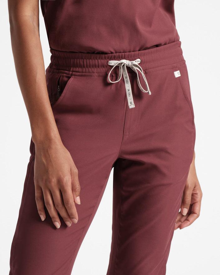 Waist to knee view of woman in straight leg deep red scrub pants with matching scrub top tucked in and branded waistband tie.