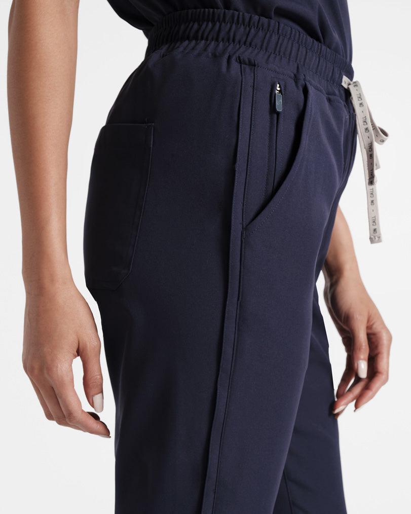 Detail view of woman in straight leg navy scrub pants, featuring the side stripe detailing, side pocket with hidden zip compartment, and branded waistband tie.