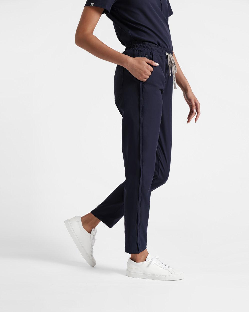 Side view of woman with hand in pocket of straight leg navy scrub pants with stripe detailing.