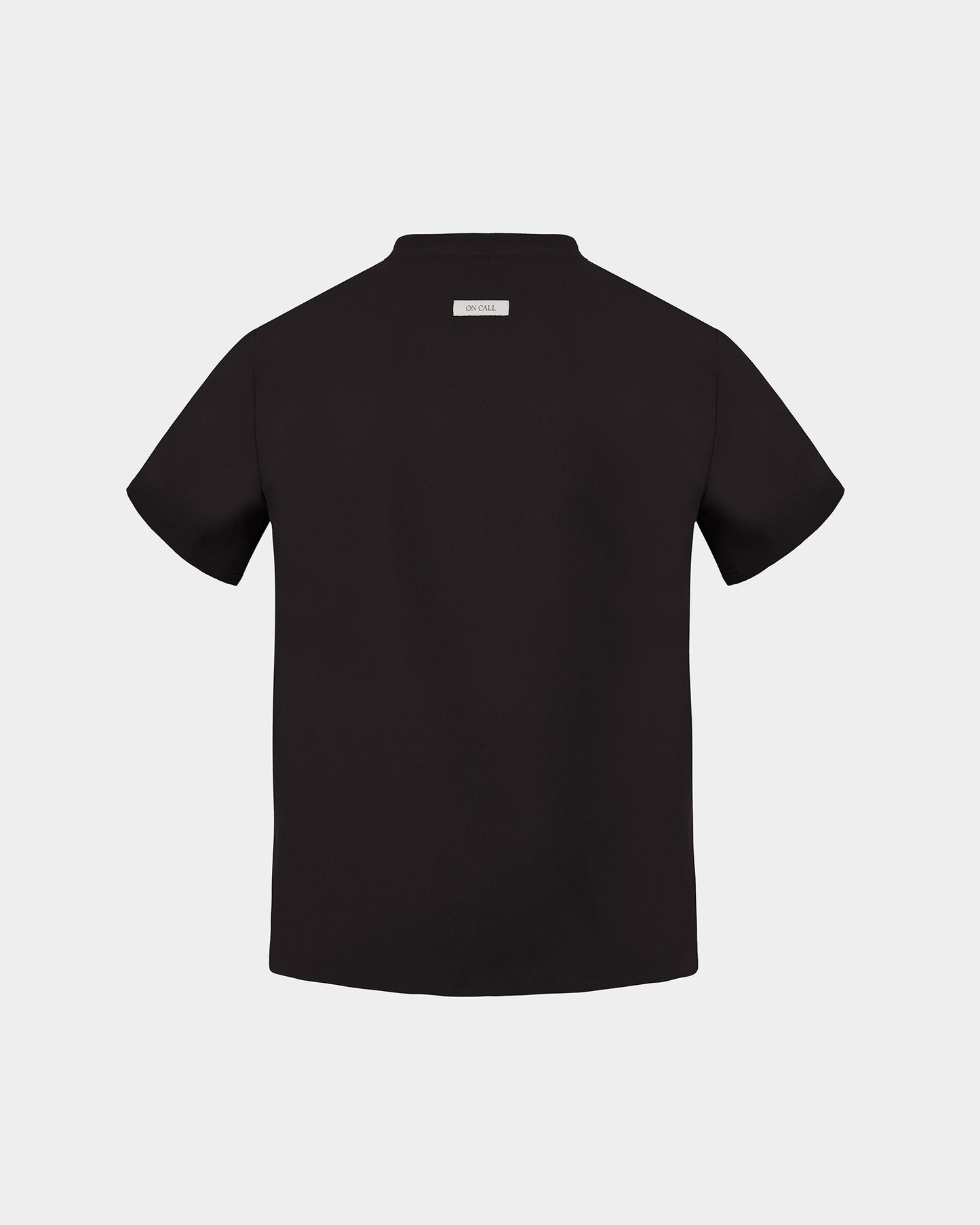 A black short-sleeve scrub shirt shown from the back, featuring a small rectangular brand label at the collar, set against a solid light gray background.