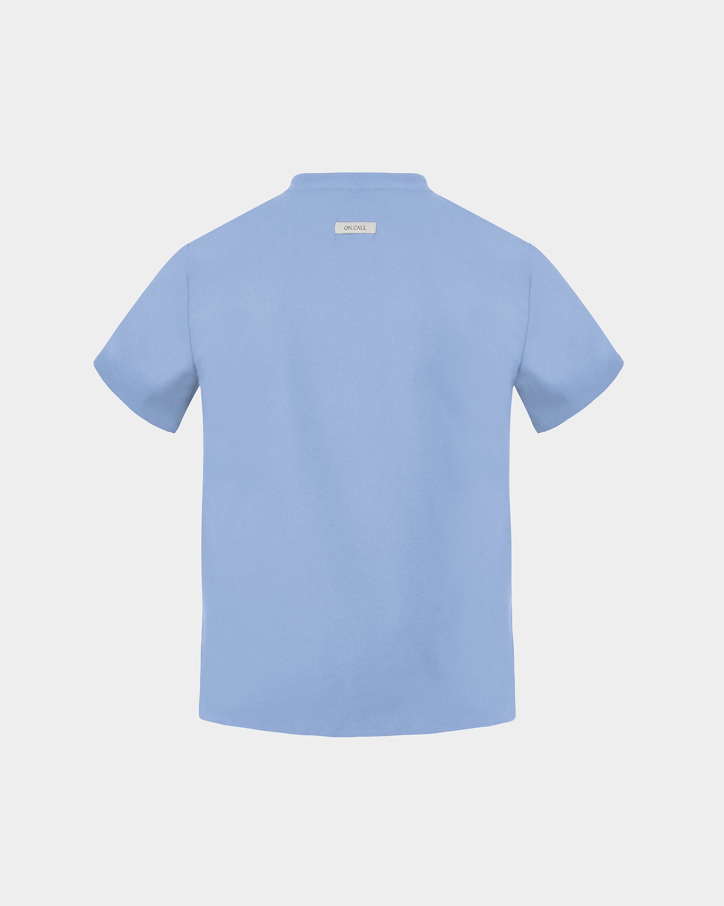A light blue short-sleeve scrub shirt shown from the back, featuring a small rectangular brand label at the inner collar, set against a solid light gray background.