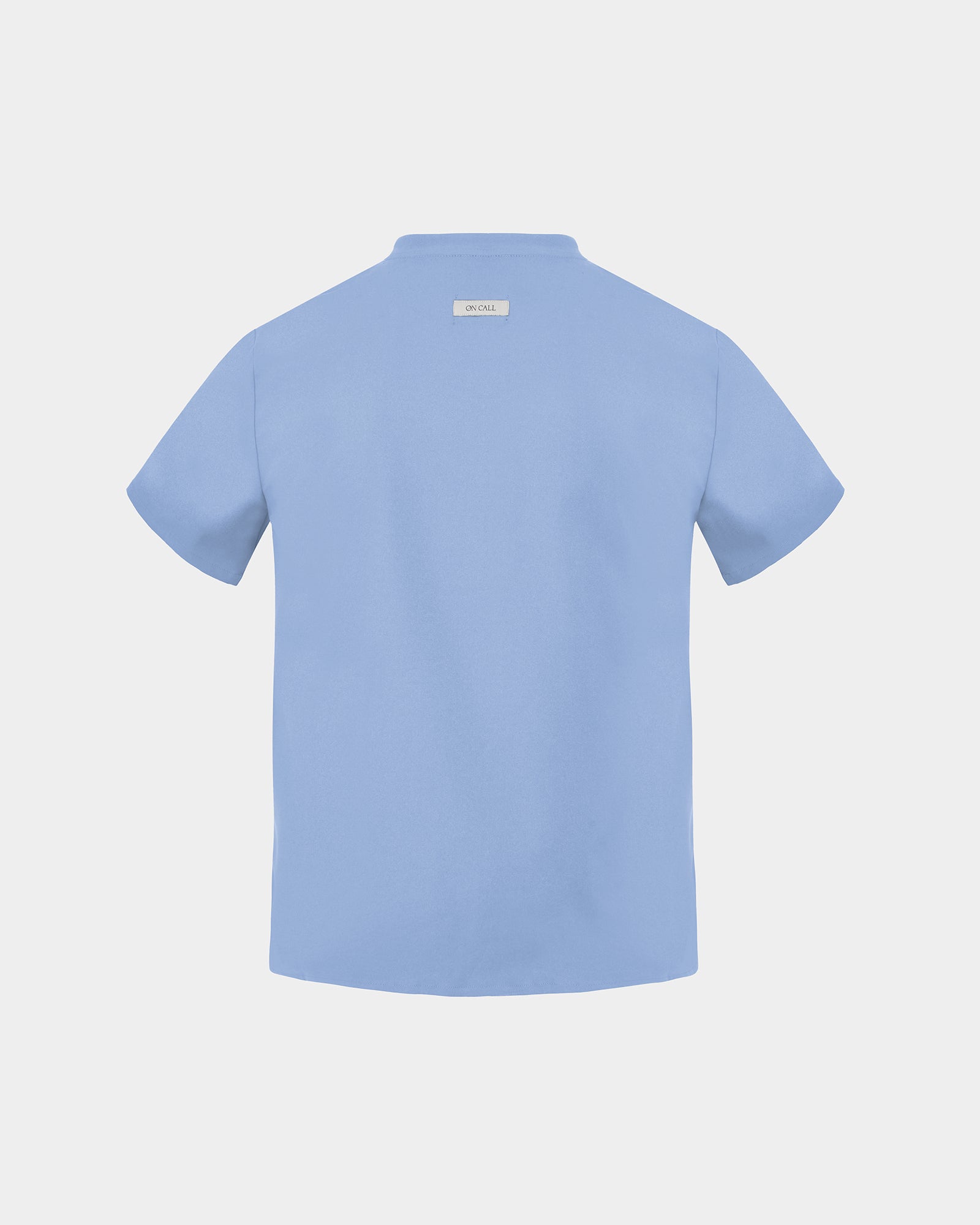 A light blue short-sleeve scrub shirt shown from the back, featuring a small rectangular brand label at the inner collar, set against a solid light gray background.