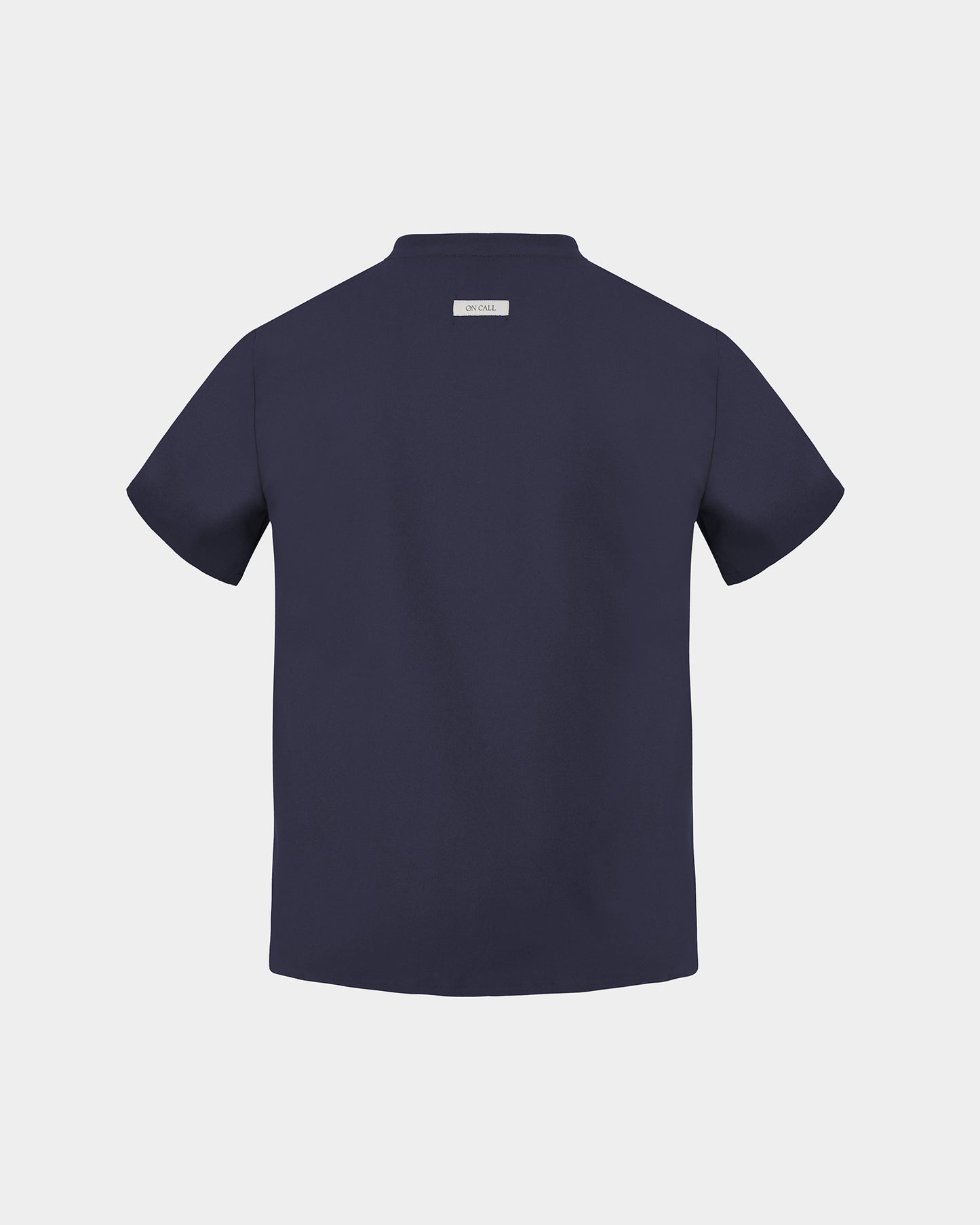 A navy short-sleeve scrub shirt shown from the back, featuring a small rectangular brand label at the collar, set against a solid light gray background.