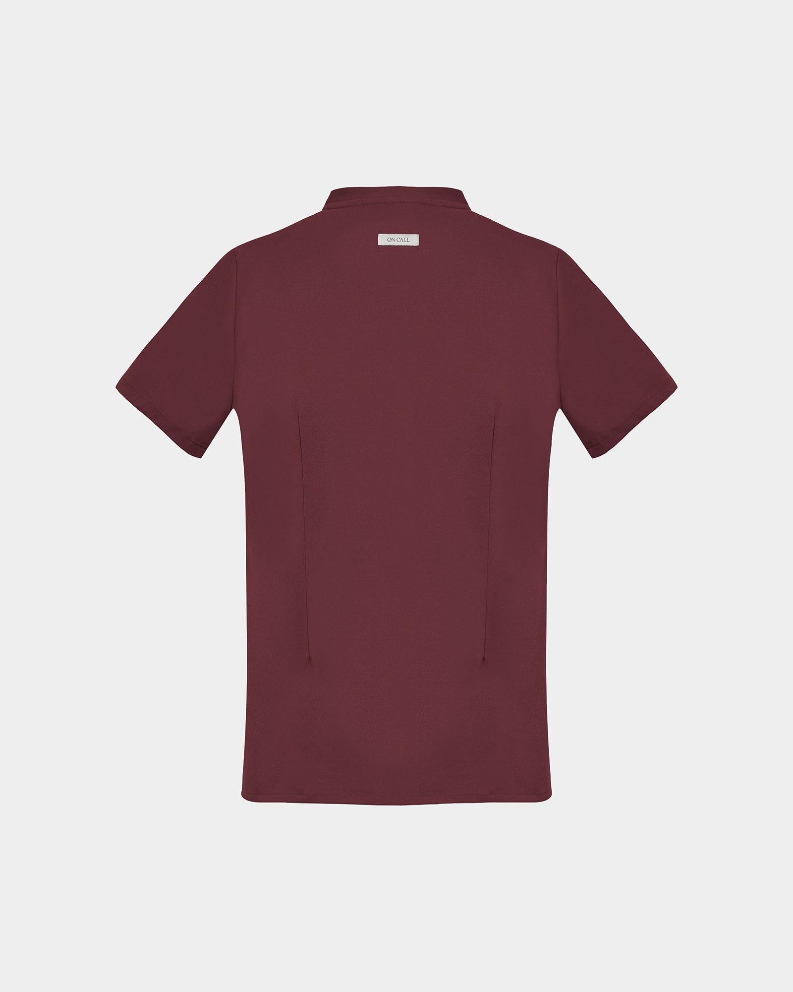 A short-sleeve scrub shirt in a deep heme red color, shown from the back, featuring two back darts, against a light gray background