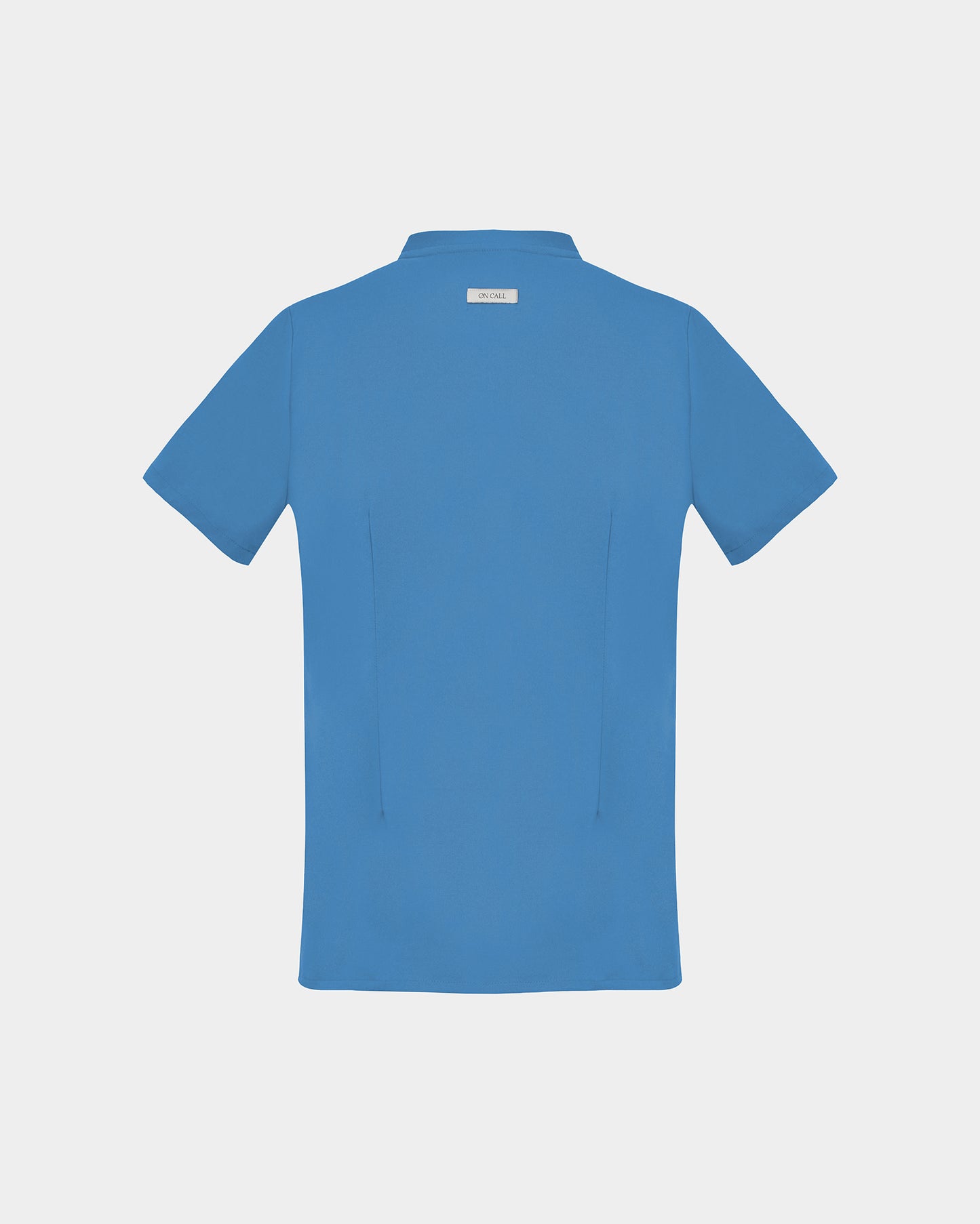 A short-sleeve scrub shirt in a solid medium blue color, shown from the back, featuring two darts, on a light gray background.