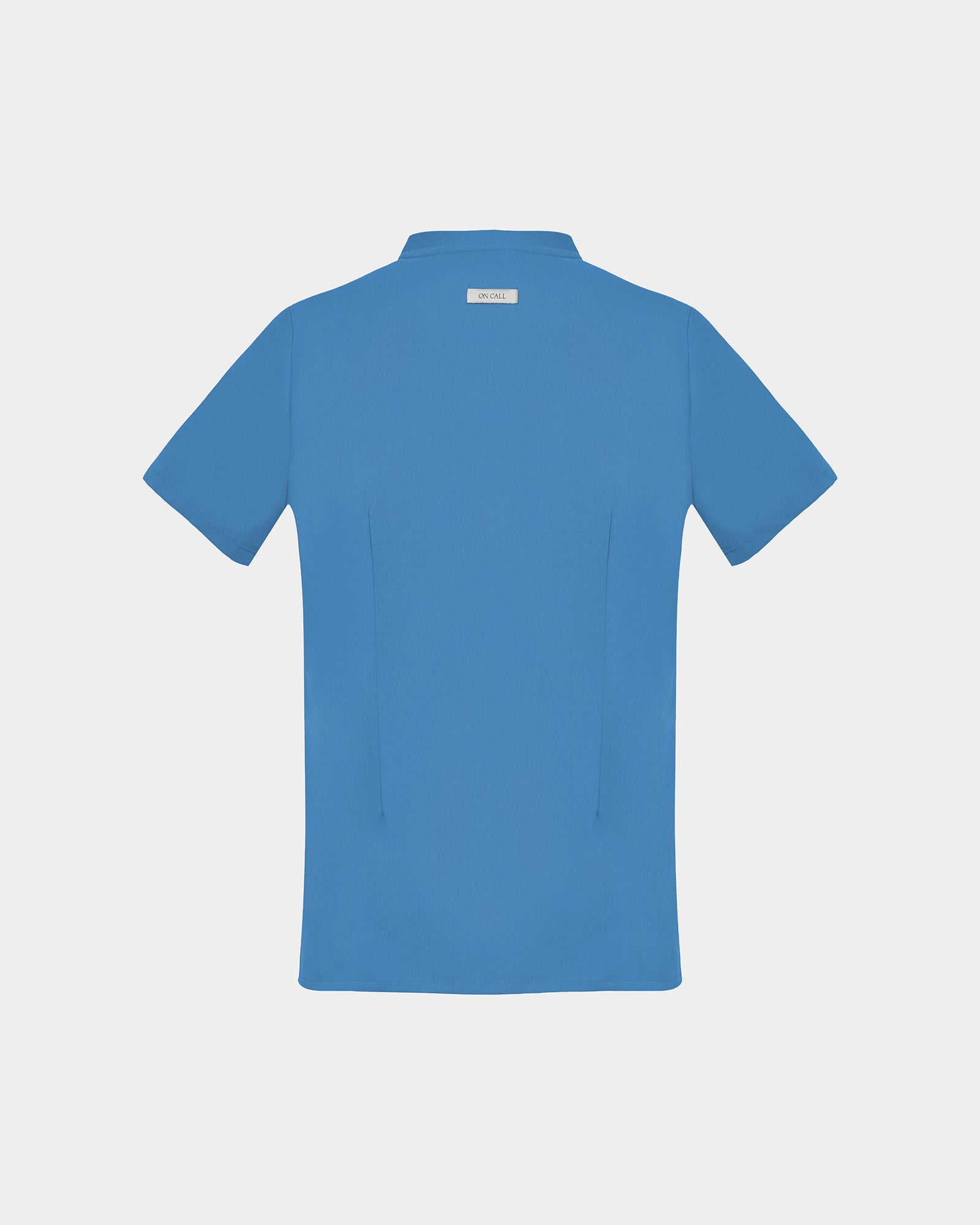 A short-sleeve scrub shirt in a solid medium blue color, shown from the back, featuring two darts, on a light gray background.