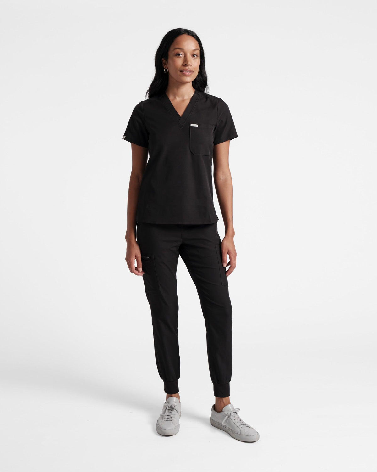 Full body view of woman wearing a black v-neck scrub top with chest pocket and matching scrub pants.