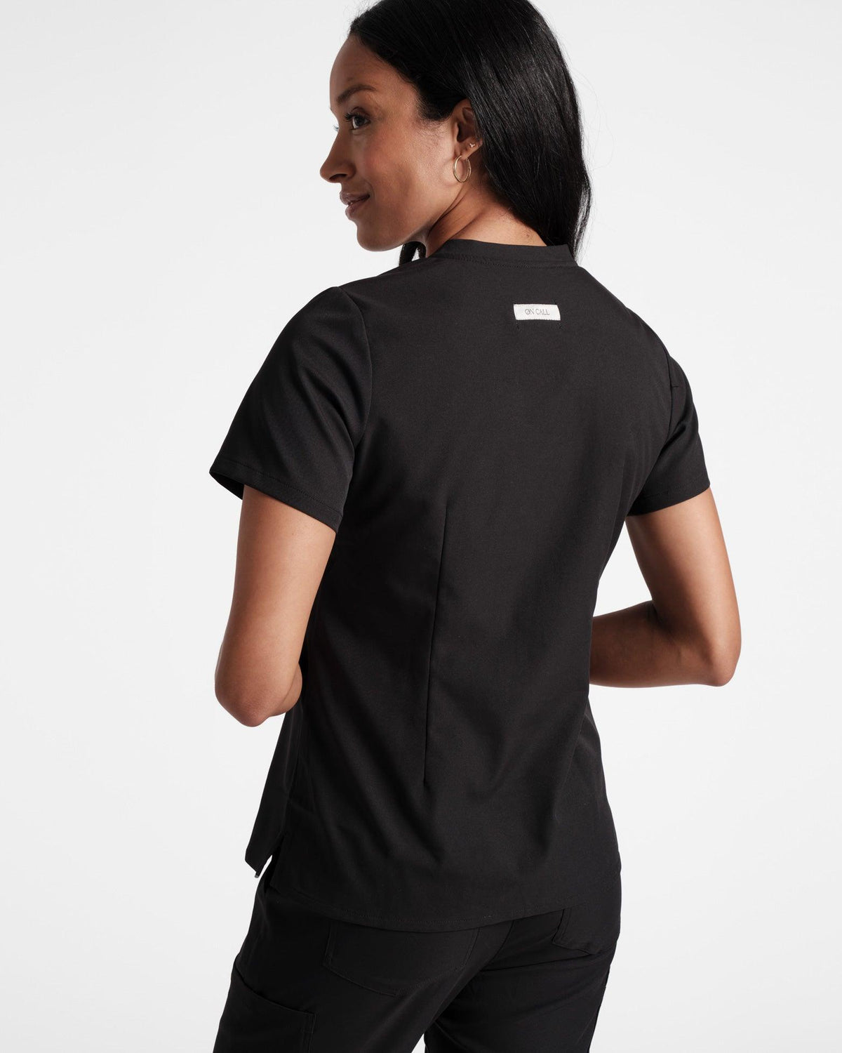 Back side view of woman wearing black scrub top with tailored dart detailing and branded fabric label under the neck seam.