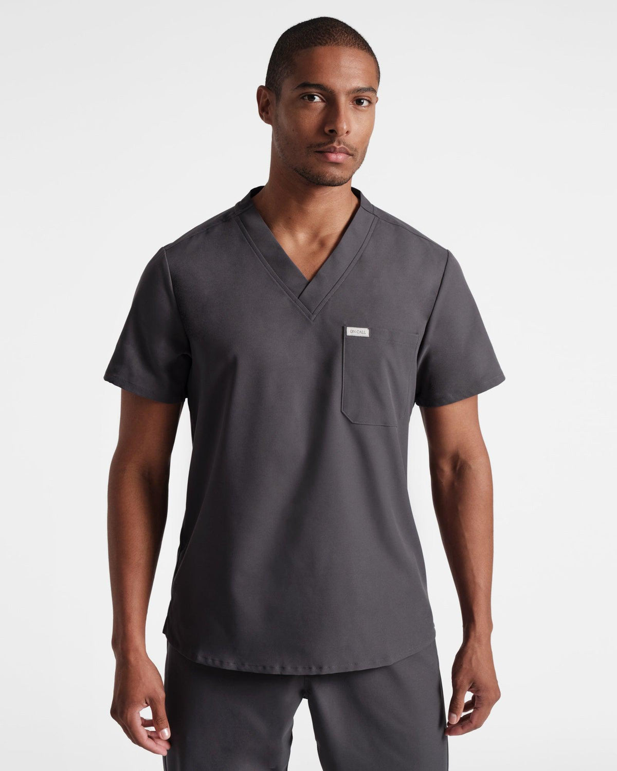 Man wearing a dark grey v-neck scrub shirt with a chest pocket, untucked over matching scrub pants.