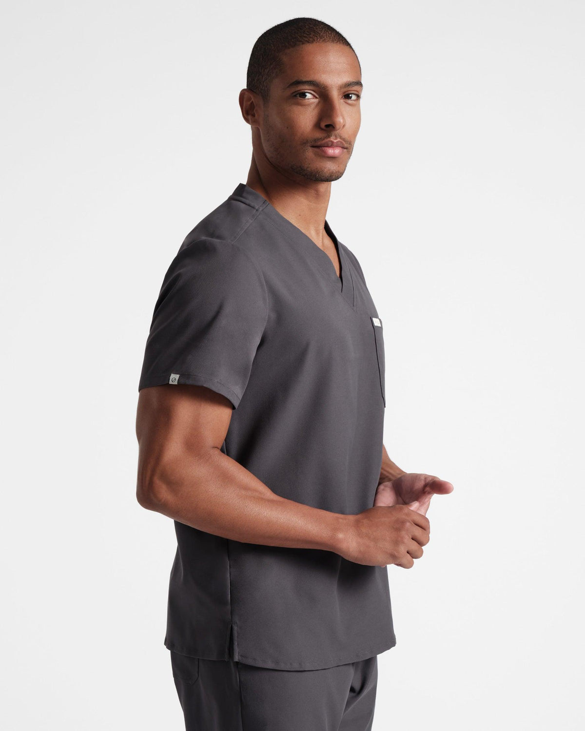 Angled side view of man in a dark grey v-neck scrub shirt, untucked over matching scrub pants.