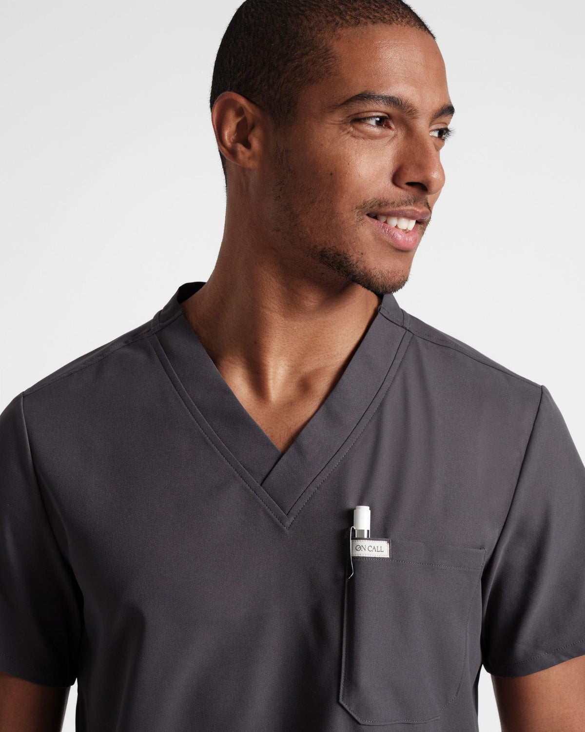 Man wearing a dark grey scrub shirt with a v-neck and pen in the chest pocket, focused on the mid-chest up.