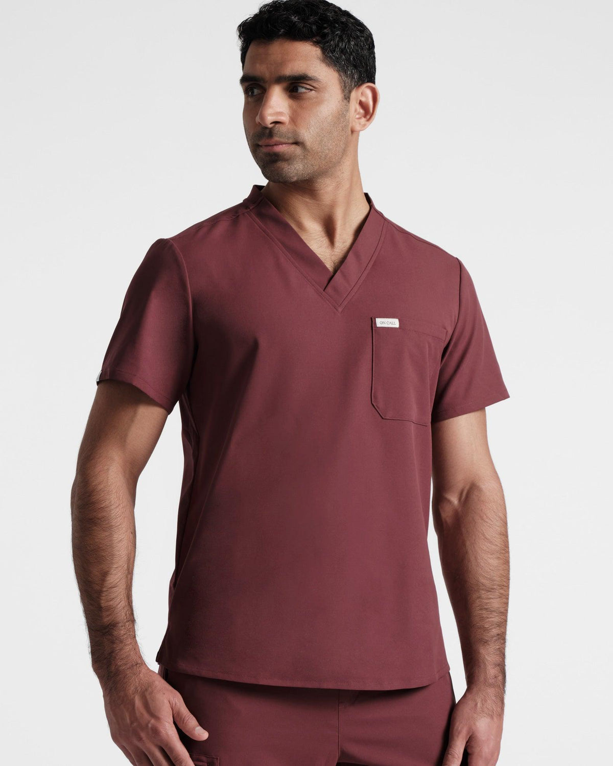 Man wearing a deep red v-neck scrub shirt with a chest pocket, looking off to the side.