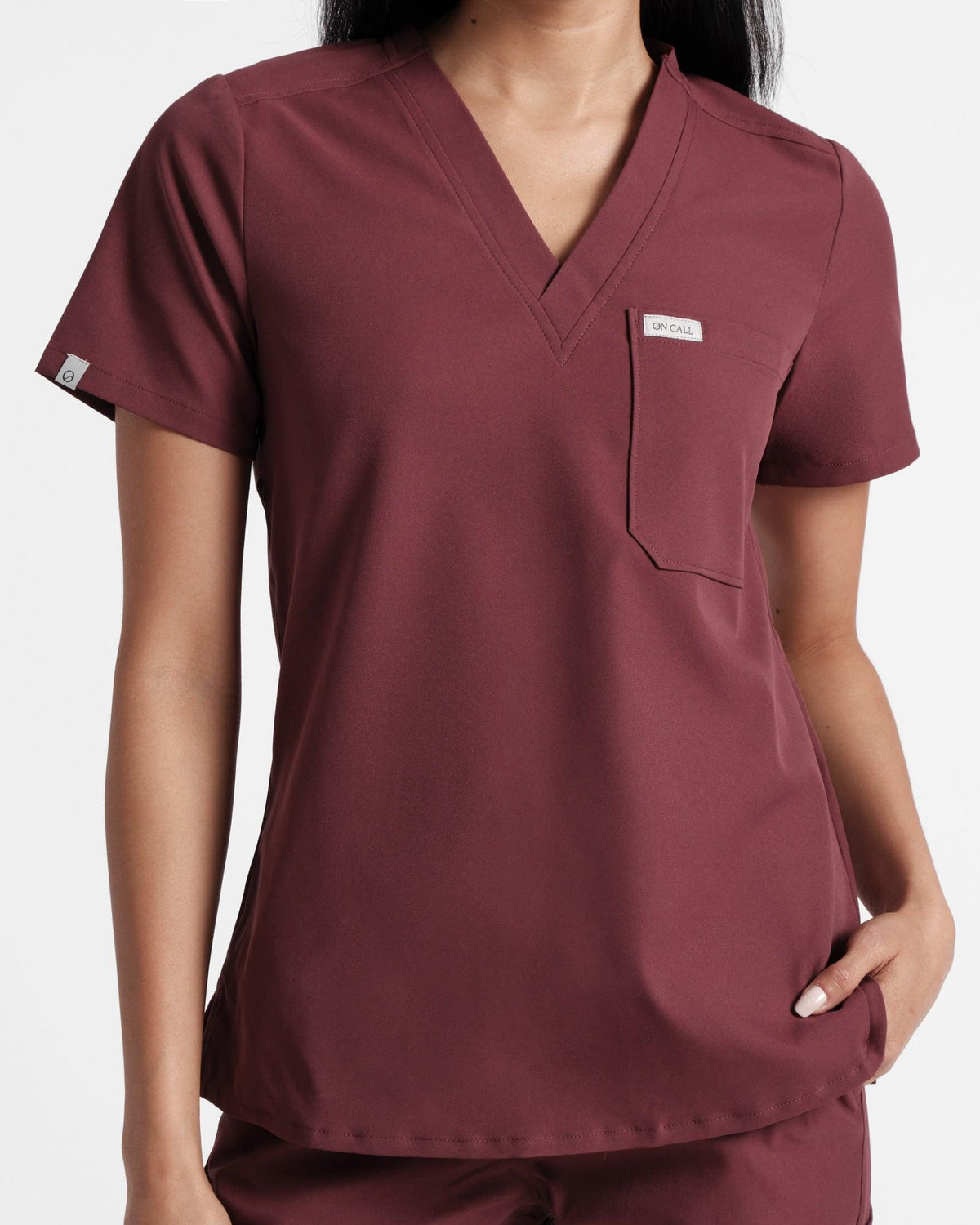 Woman in deep red scrub shirt with v-neck, pocket on the left chest, and small logo label wrapped around the right sleeve's hem. 