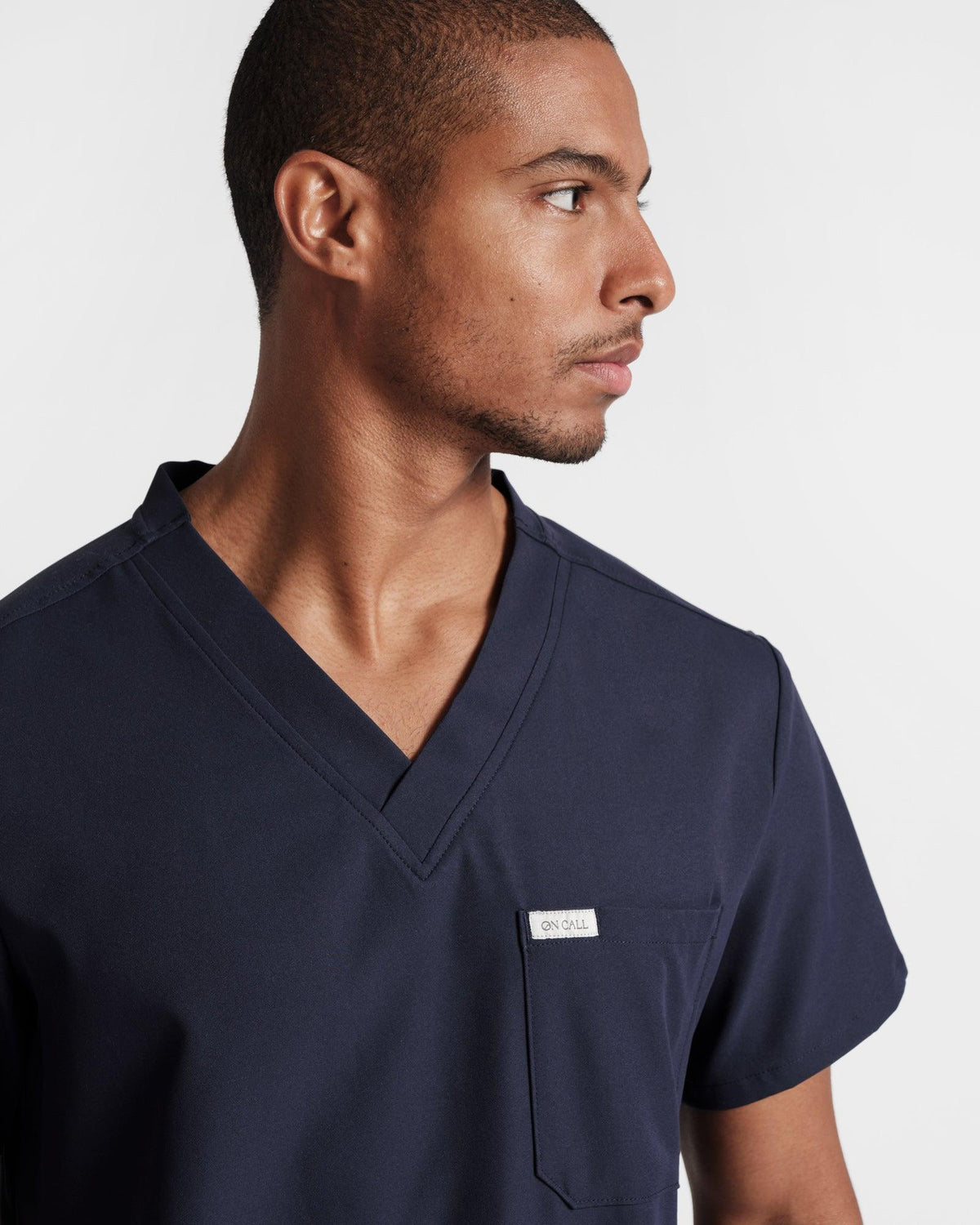 Man wearing a navy v-neck scrub shirt with a chest pocket, focused from the mid-torso up.