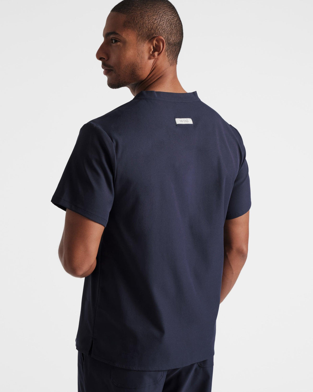 Angled back side view of man wearing a navy scrub shirt with an On Call fabric label sewn under the neck seam.