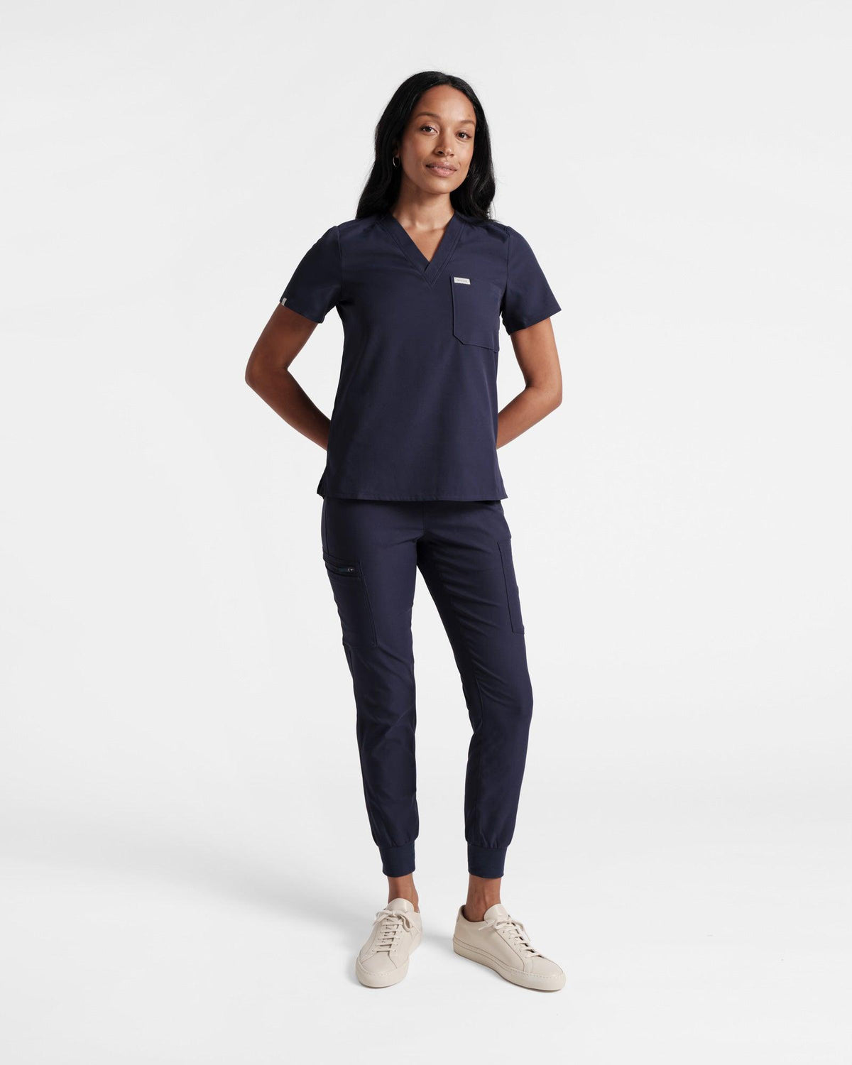 Woman in navy v-neck scrub shirt and matching jogger cargo scrub pants with her hands resting behind her back.