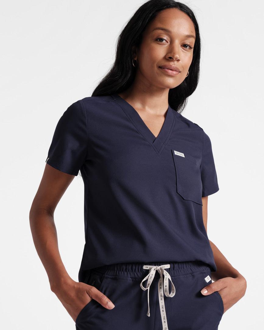 Woman in a navy v-neck scrub shirt tucked into matching scrub pants with a branded On Call waistband tie.