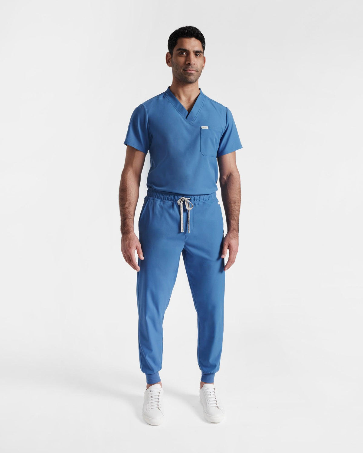 Man wearing a v-neck scrub shirt tucked into jogger scrub pants, both in a bright blue hue.
