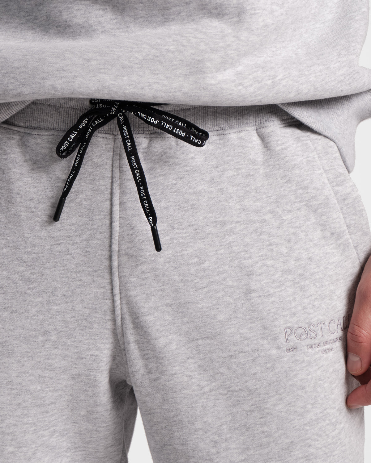 Zoomed in view of men's heather grey jogger sweatpant's branded waistband tie in black and tonal Post Call logo embroidery on the thigh.