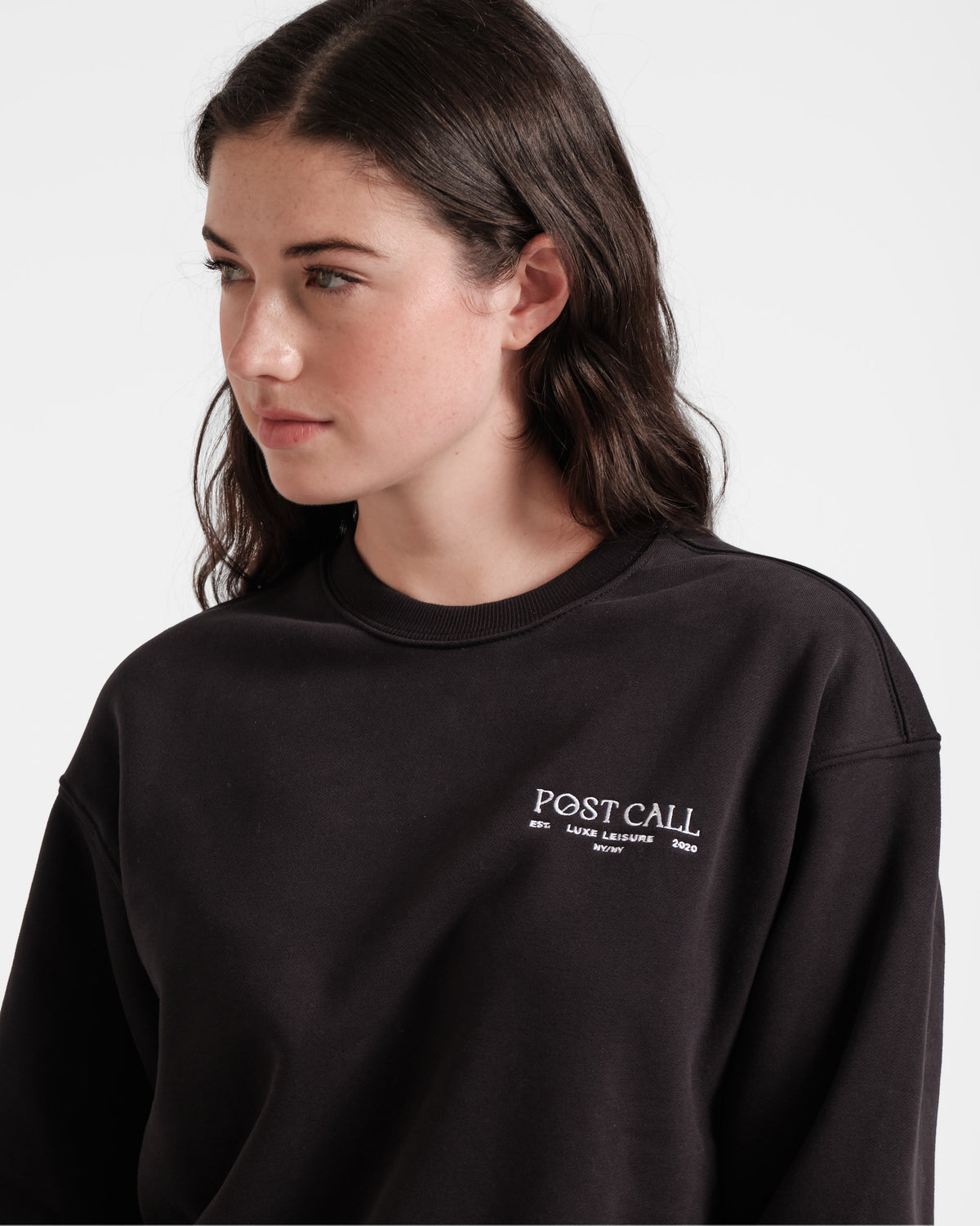 Close up view of woman wearing a unisex black crewneck sweatshirt with white Post Call logo embroidery.