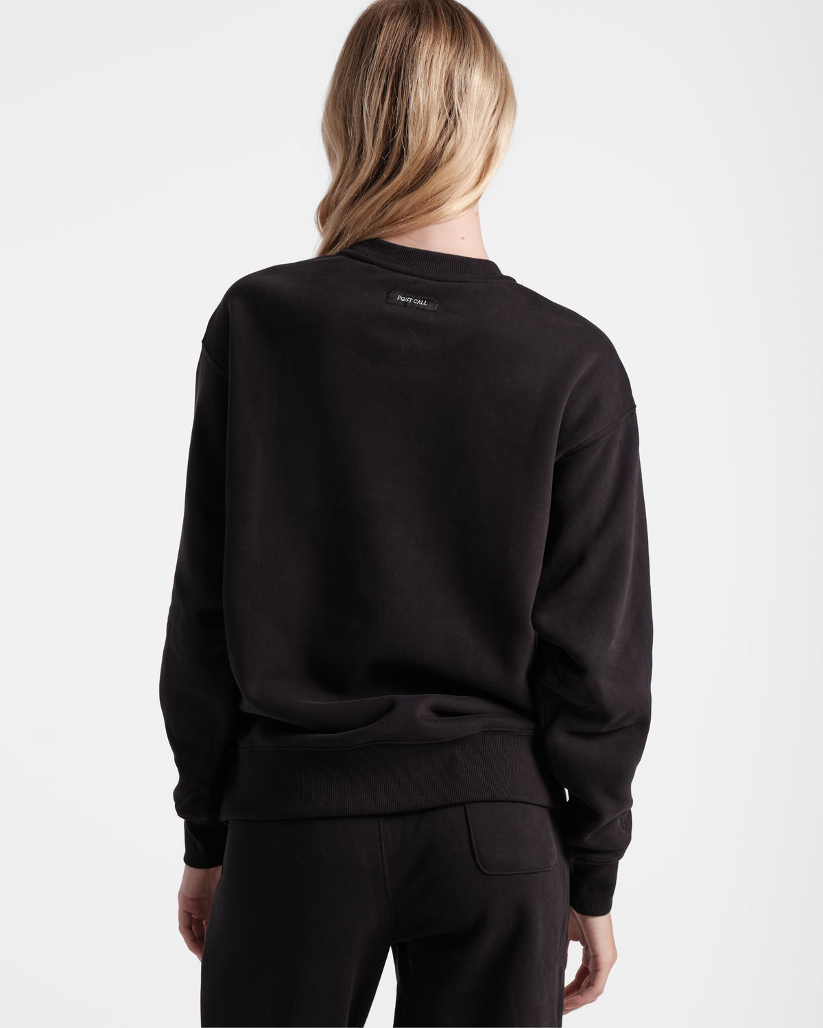 Back side view of woman wearing black crewneck sweatshirt with Post Call fabric label sewn under neck seam.