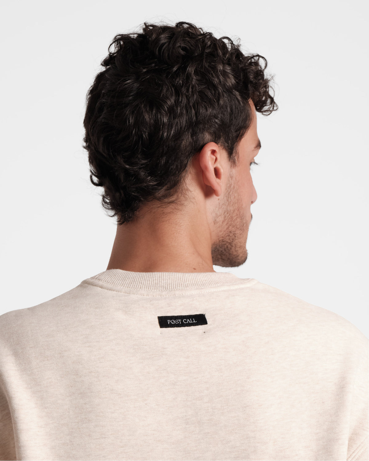 Close up of man's upper back wearing a heather beige crewneck sweatshirt with a black Post Call fabric label sewn under the neck seam.