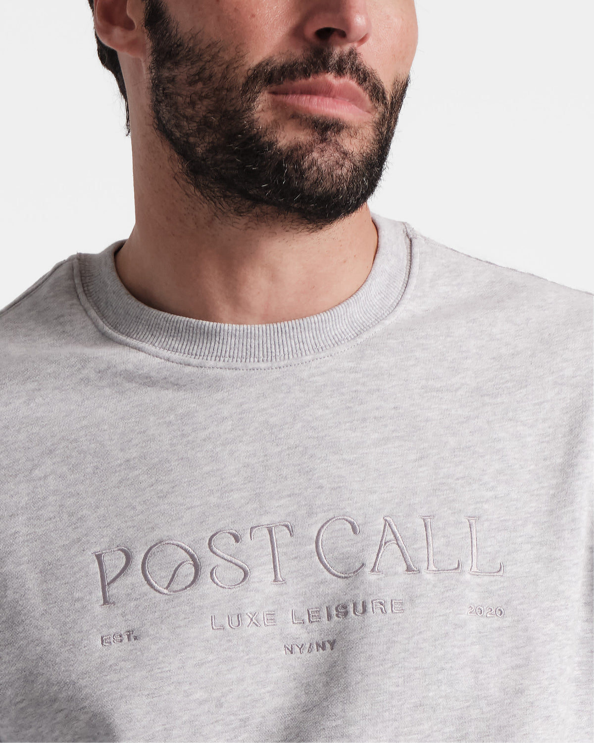 Close up from mid chest to nose of a man wearing heather grey crewneck sweatshirt with tonal Post Call logo embroidery across the chest and ribbed neck.