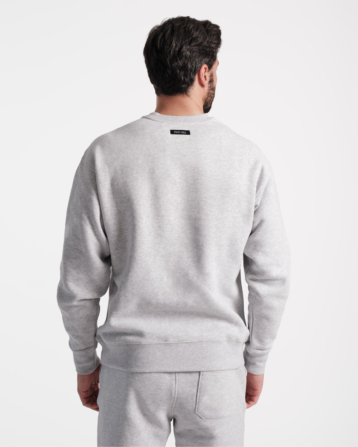 Back side view of man wearing unisex heather grey crewneck sweatshirt with black Post Call fabric label sewn under neck seam.