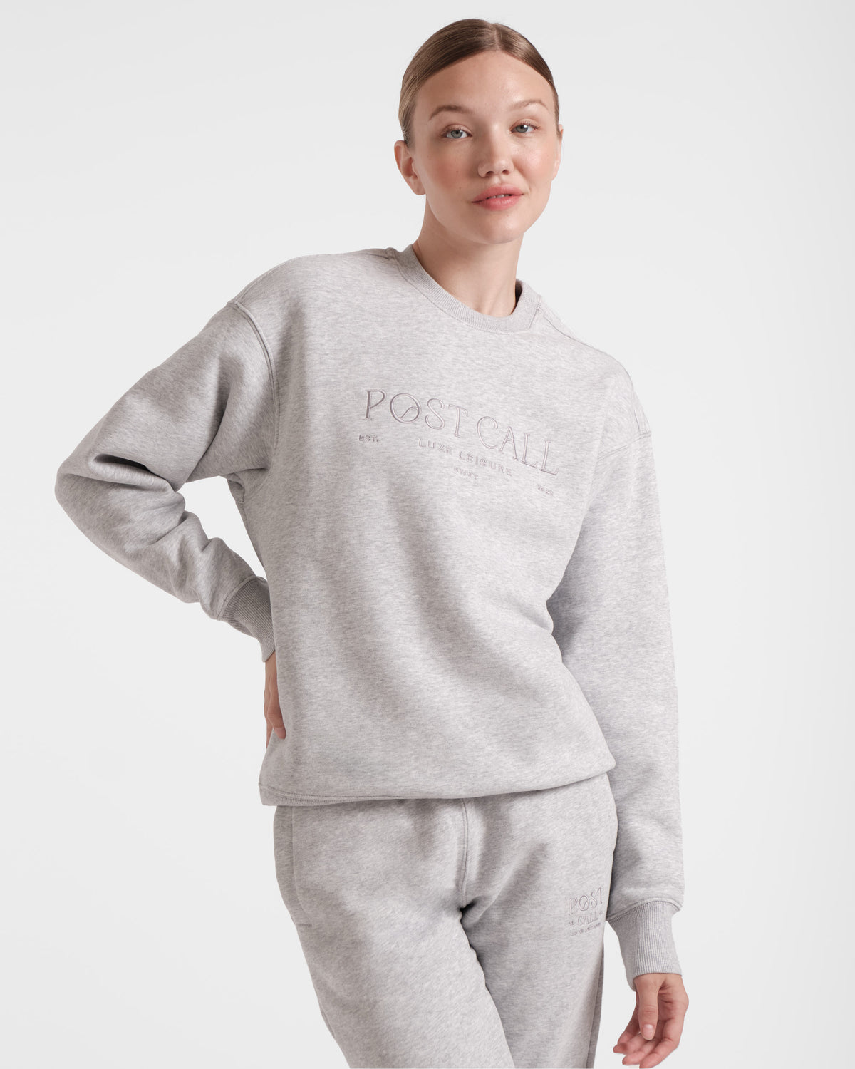 A woman wearing a heather grey crewneck sweatshirt with tonal Post Call logo embroidery and ribbed sleeve cuffs, standing with one hand on her hip.