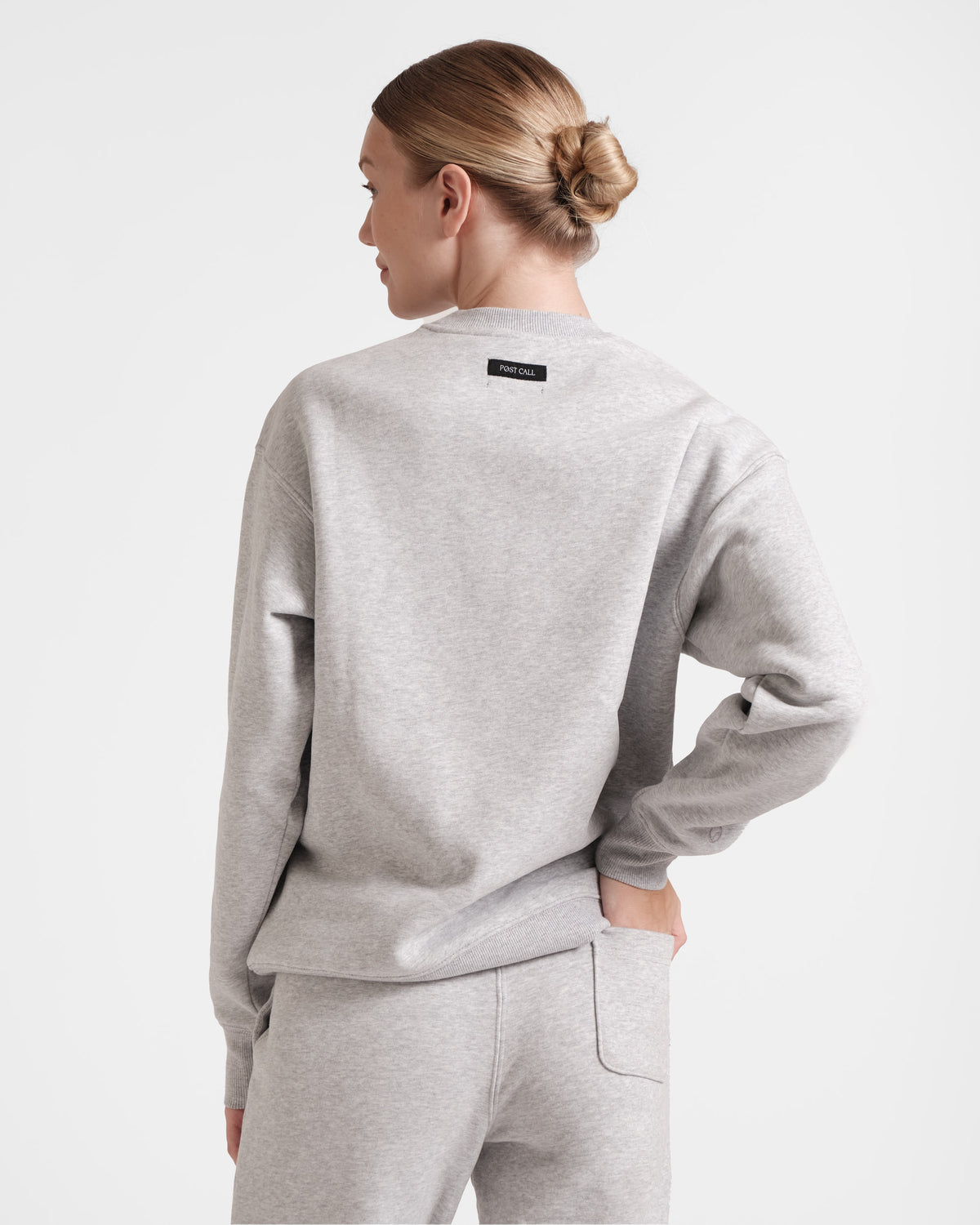 Back side view of woman wearing heather grey crewneck sweatshirt with black Post Call fabric label sewn under neck seam.