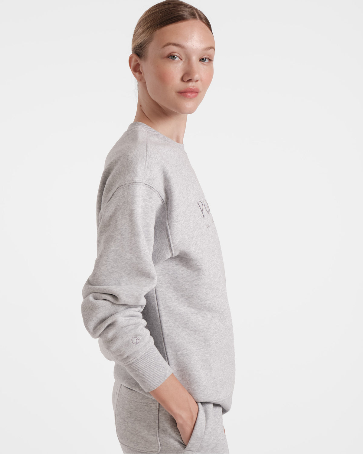 Side view of woman wearing heather grey crewneck sweatshirt with tonal Post Call logo embroidery on chest.