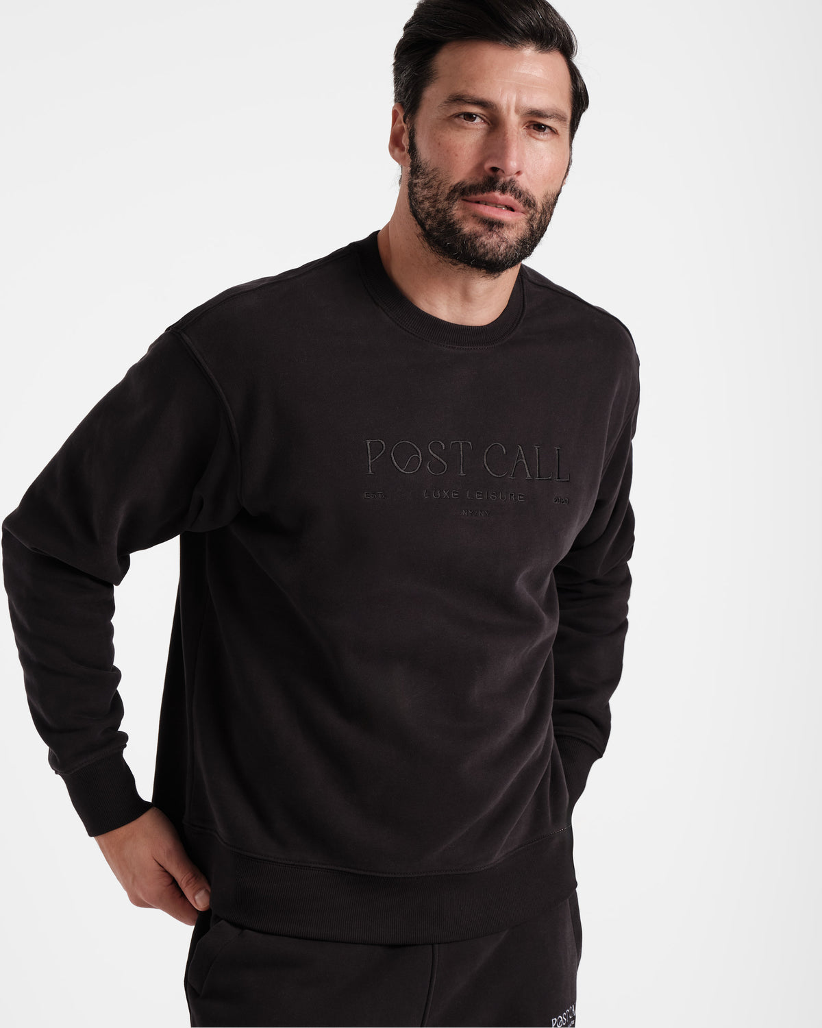 Man wearing unisex black crewneck sweatshirt with tonal Post Call logo embroidery across the chest.