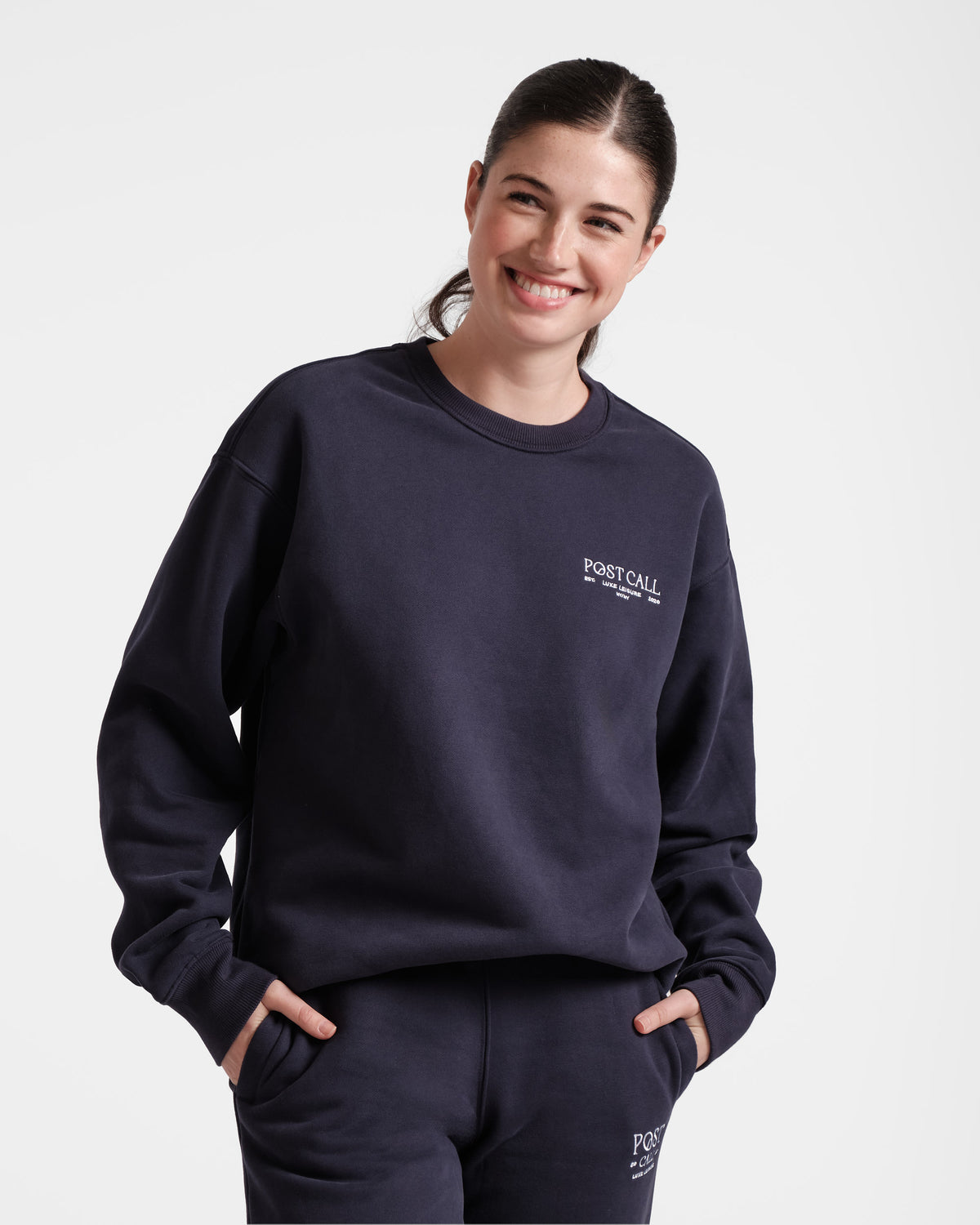 Woman wearing a navy unisex crewneck sweatshirt with white Post Call logo embroidery on the left chest.