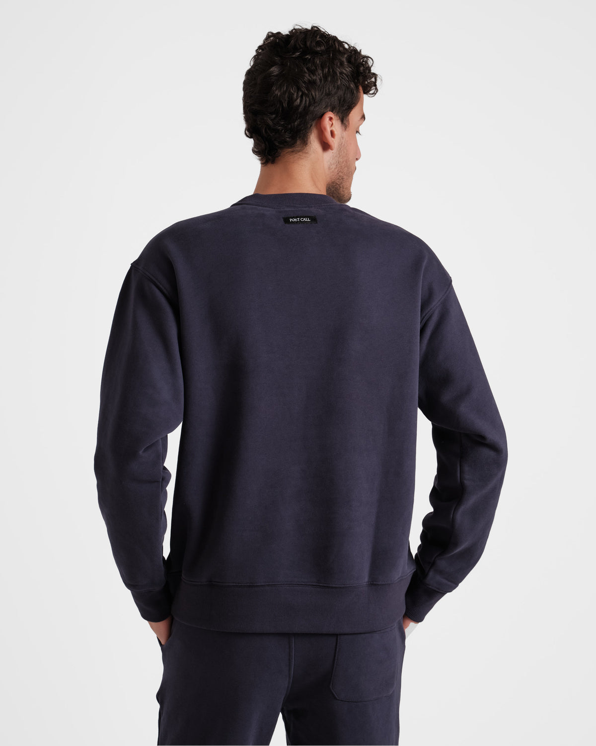 Back side view of man wearing unisex navy crewneck sweatshirt with black Post Call fabric label sewn under neck seam.