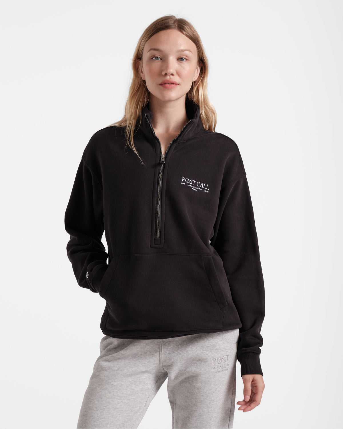 Woman wearing a men's cut black half zip sweatshirt with a kangaroo pocket and white Post Call logo embroidered on left chest.