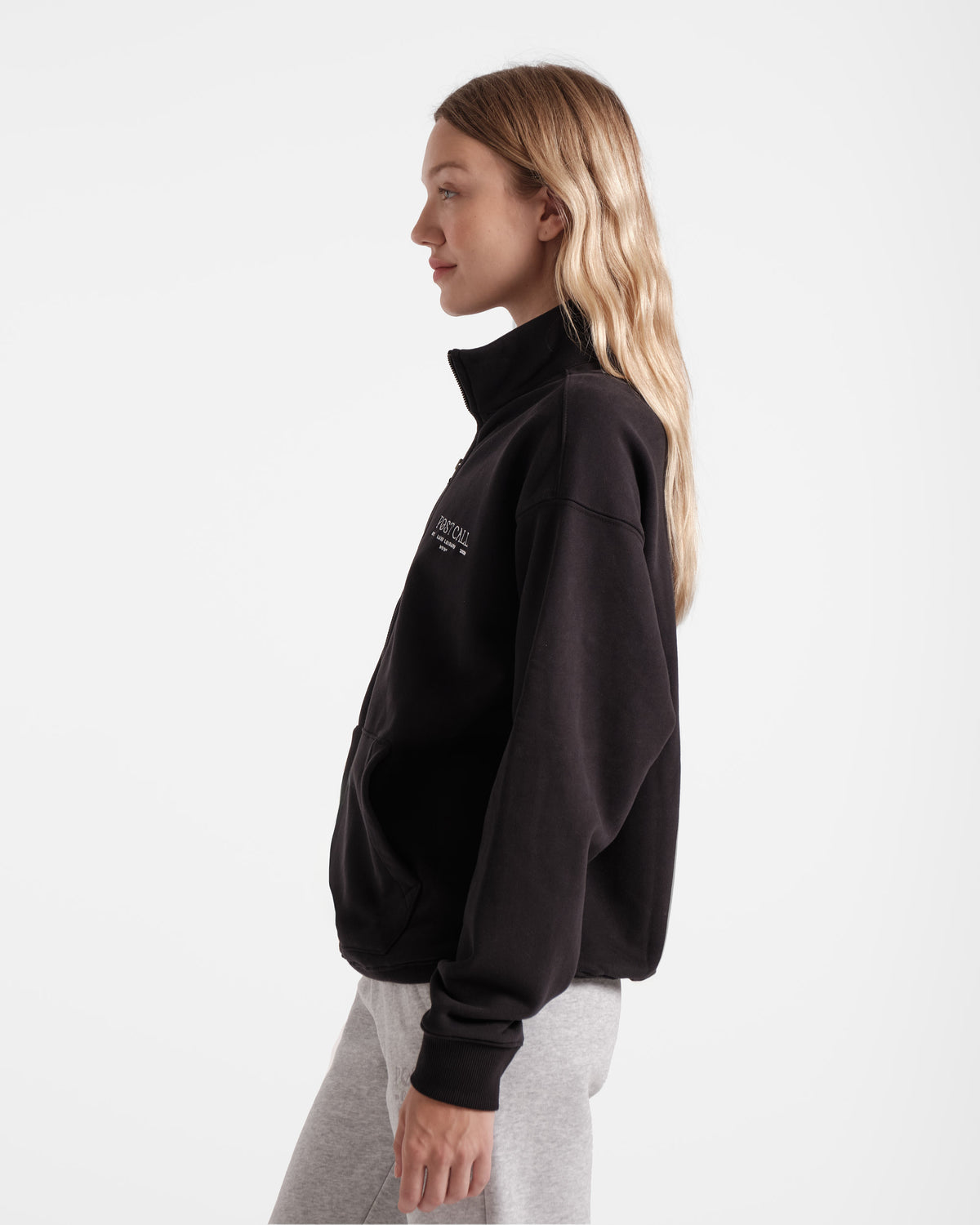 Side view of woman wearing a men's cut black half zip sweatshirt with a kangaroo pocket and white Post Call logo embroidered on the left chest.