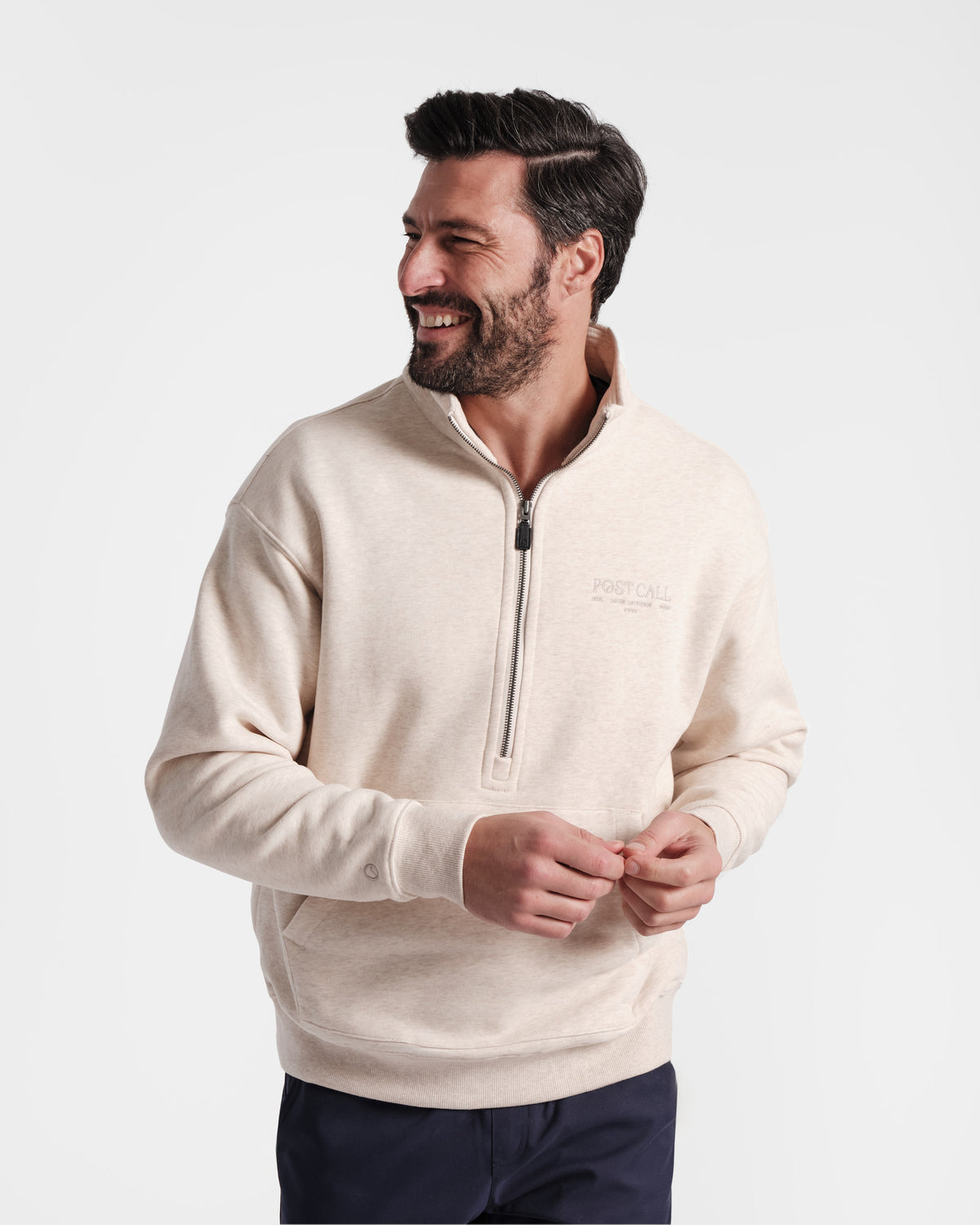Man wearing heather beige half zip sweatshirt with a kangaroo pocket and tonal Post Call logo embroidered on the left chest.